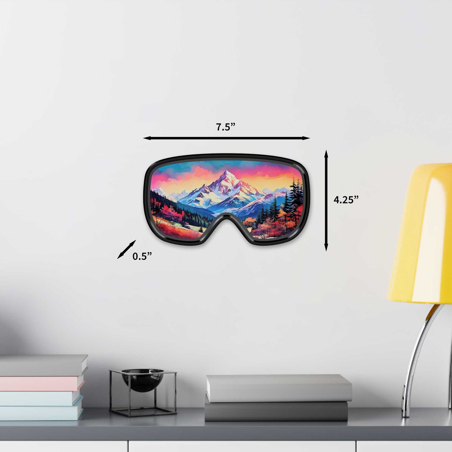 Decorative ski goggle wall art measuring 7.5 inches wide by 4.25 inches tall, showcasing a vivid pink, orange, and purple mountain landscape, displayed on a white wall next to a modern yellow lamp.