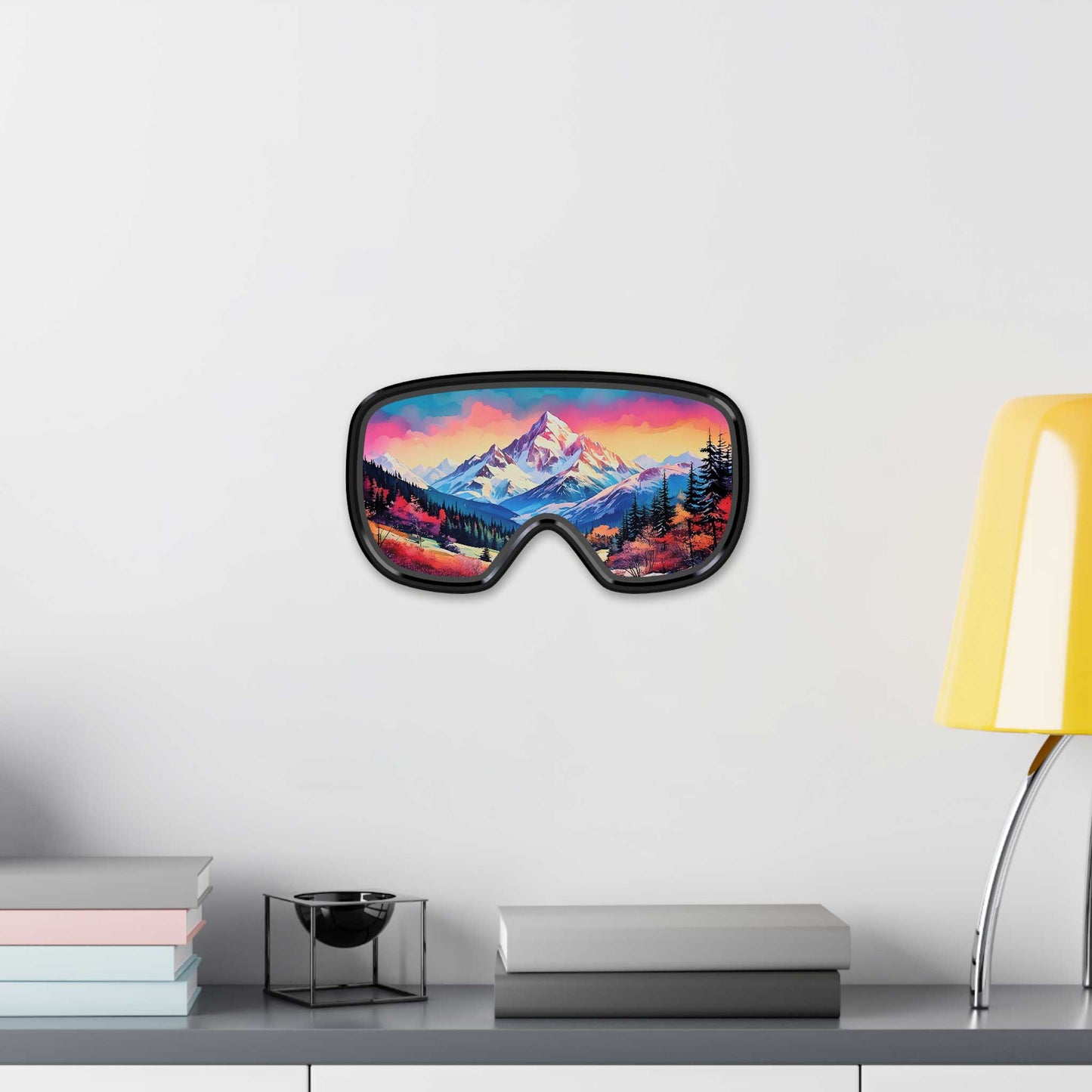 Vibrant ski goggle wall art featuring a bold pink, orange, and purple mountain landscape, hanging on a white wall next to a modern yellow lamp and stacked books.